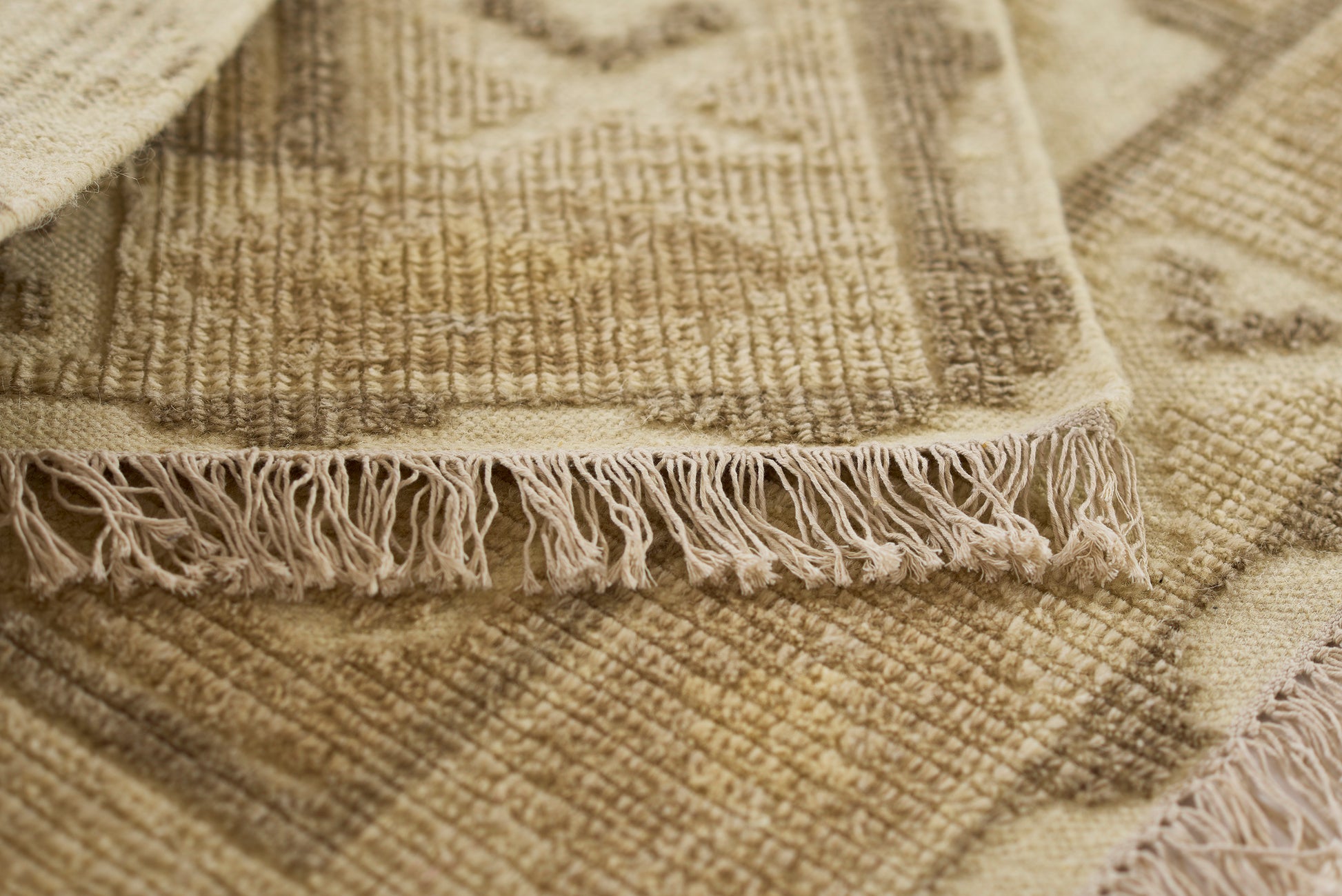 Beige woven rug or throw with fringed edges, textured geometric pattern in warm neutral tones.