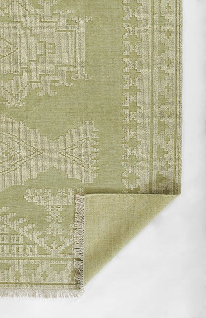 Olive green geometric woven rug with beige pattern, fringed edge, folded corner.