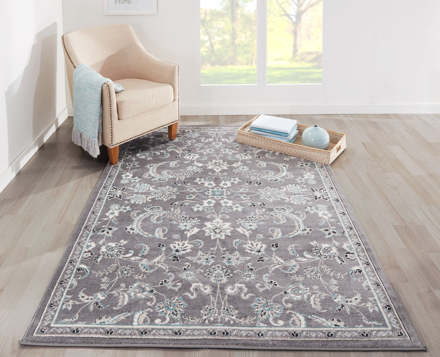Gray traditional area rug with teal floral motifs, beige upholstered armchair, light wood floor, wicker tray with books.