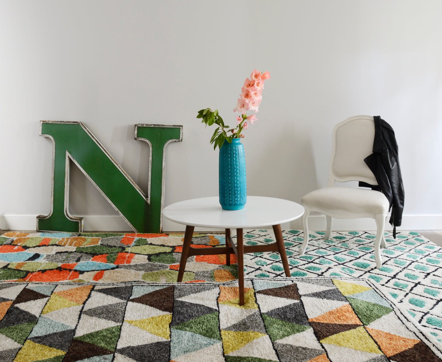 Teal vase with pink flowers on white round table, green letter N, white upholstered chair, black jacket, geometric rug.