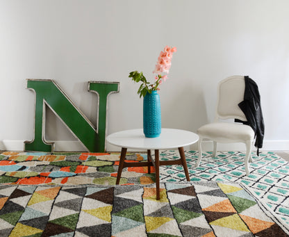 Teal vase with pink flowers on white round table, green letter N, white upholstered chair, black jacket, geometric rug.