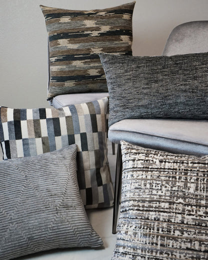 Decorative throw pillows in gray, taupe, and brown with geometric, striped, and woven textures, stacked near a gray chair.