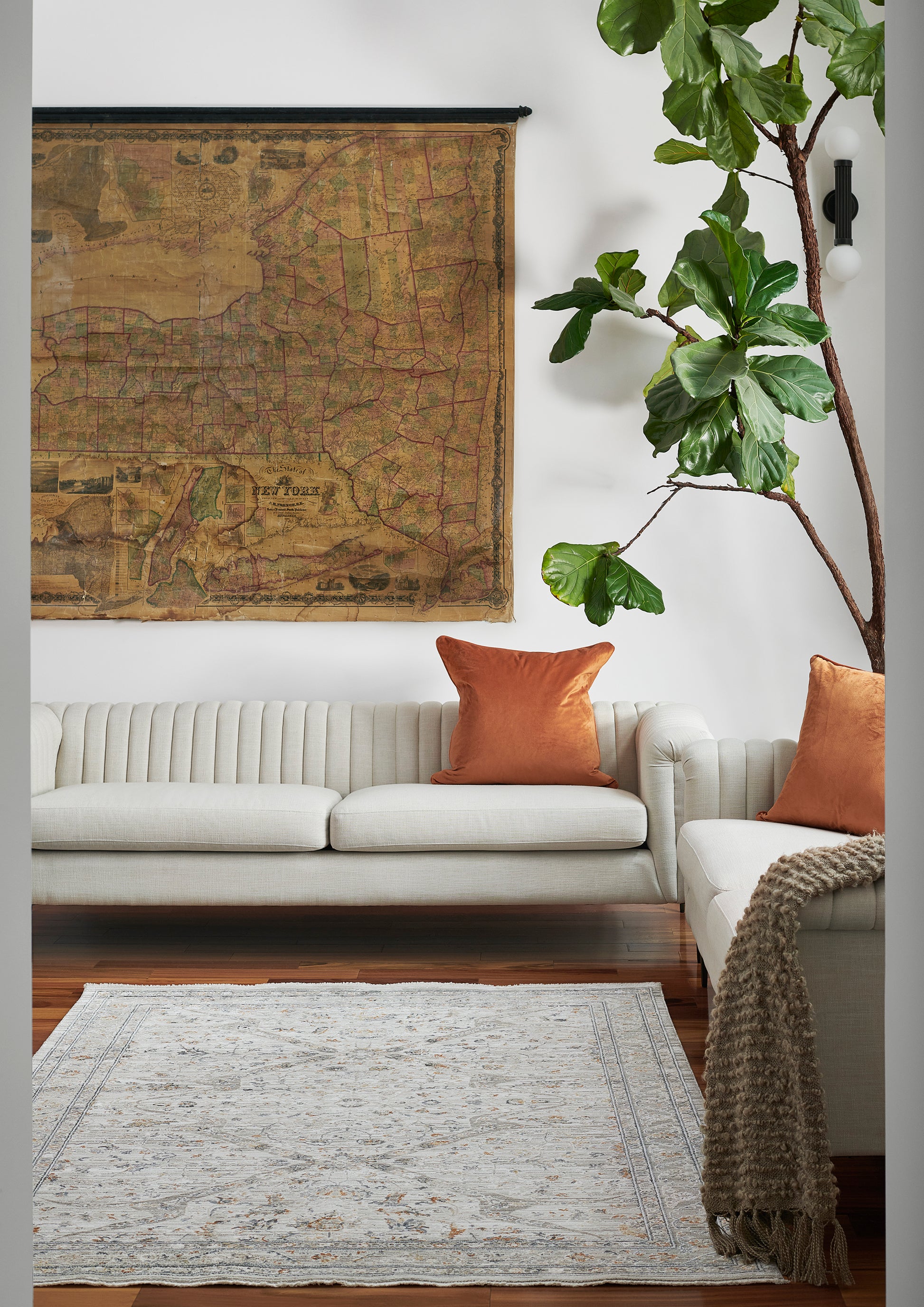Living room with large vintage map wall art, cream sectional sofa, orange pillows, pale rug, and tall fiddle-leaf fig.