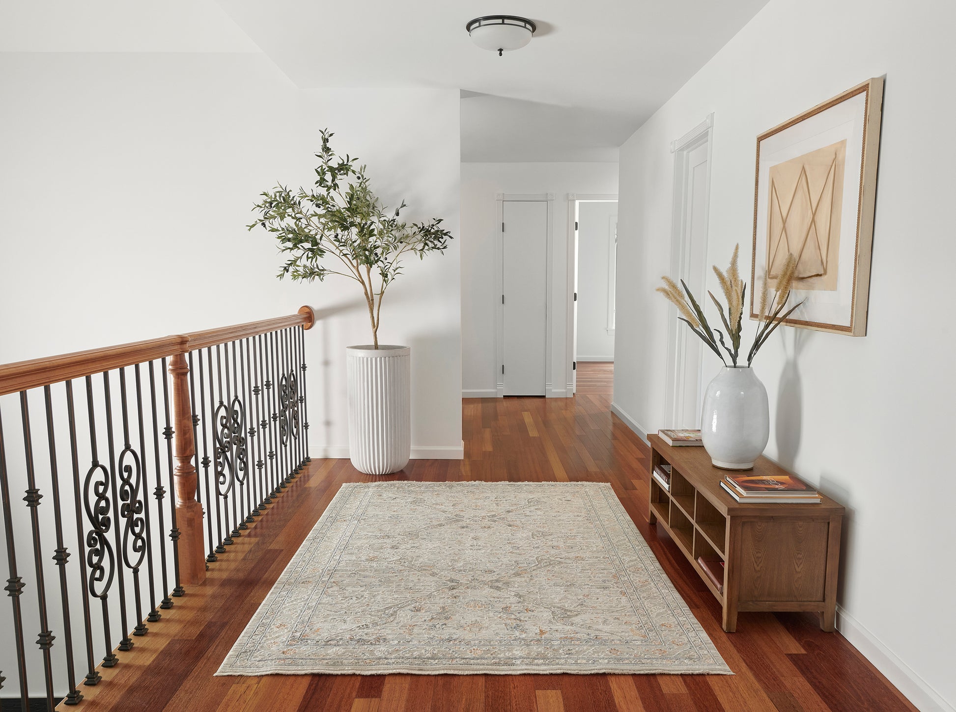 Bright hallway with wood floor, black iron railing, tall potted plant, light patterned rug, and wooden console with vase.