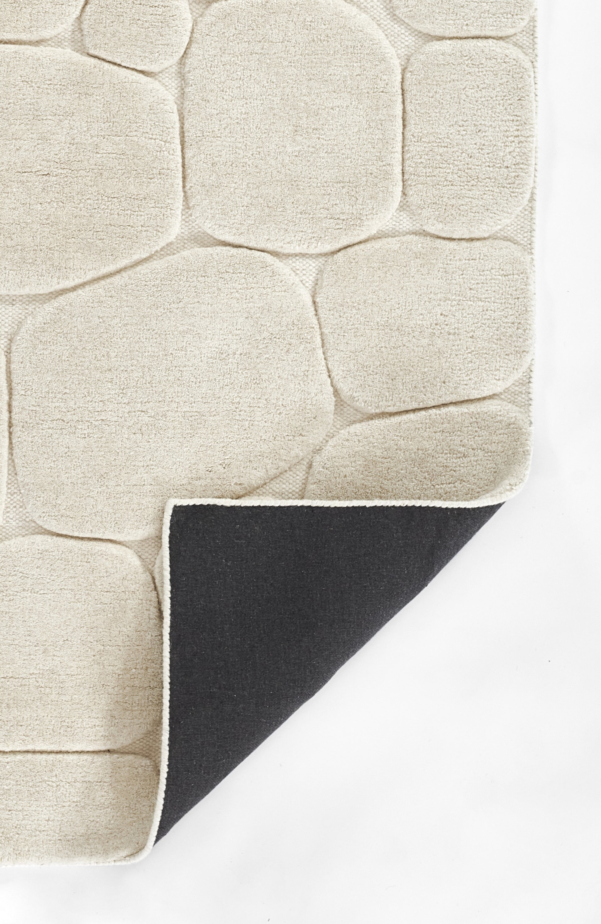 Cream tufted rug with pebble-like blocks and soft pile; folded corner reveals black underside and white edge.