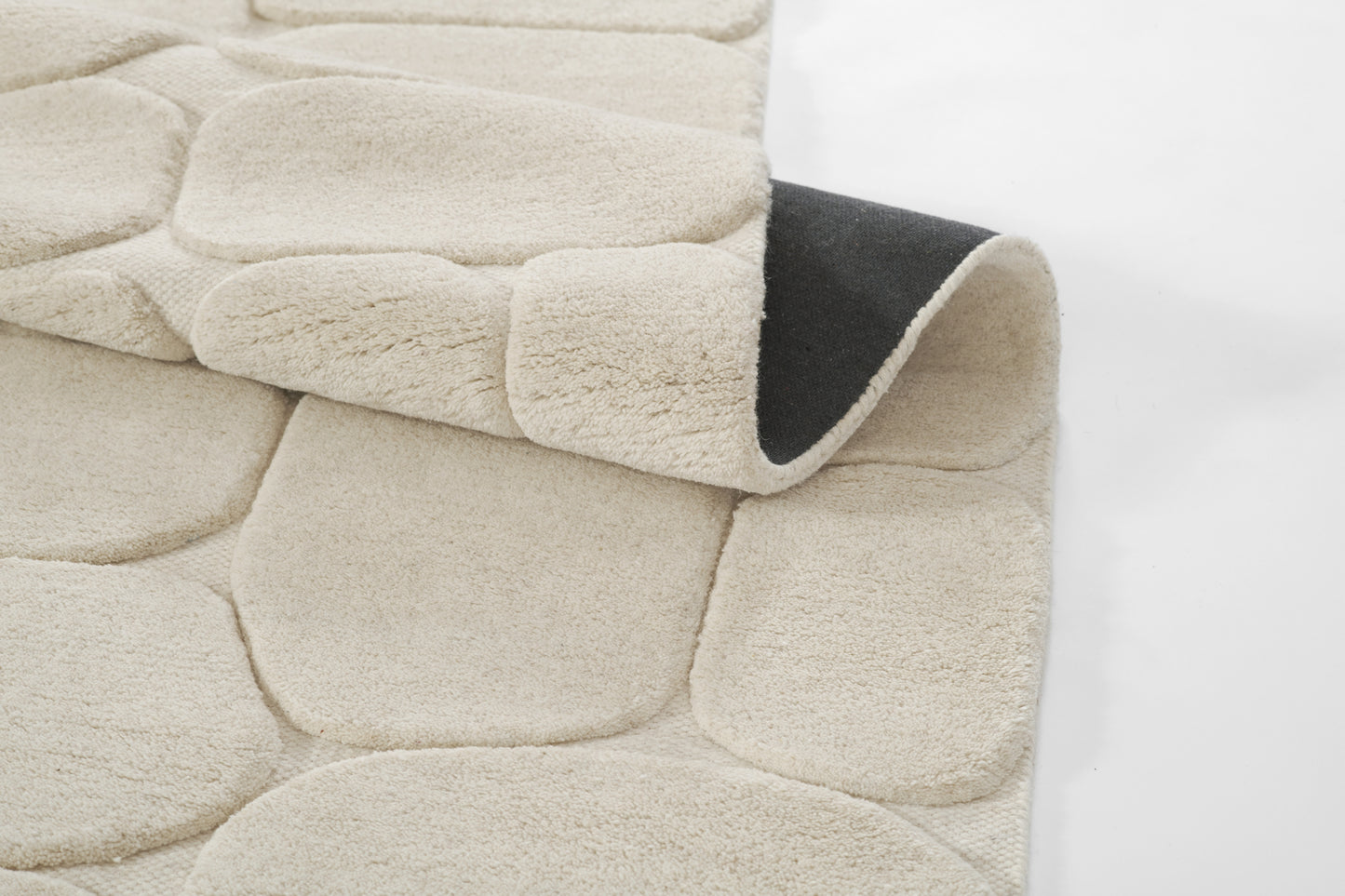Beige plush pebble rug with rounded stone-like panels; edge rolled to reveal dark underside.