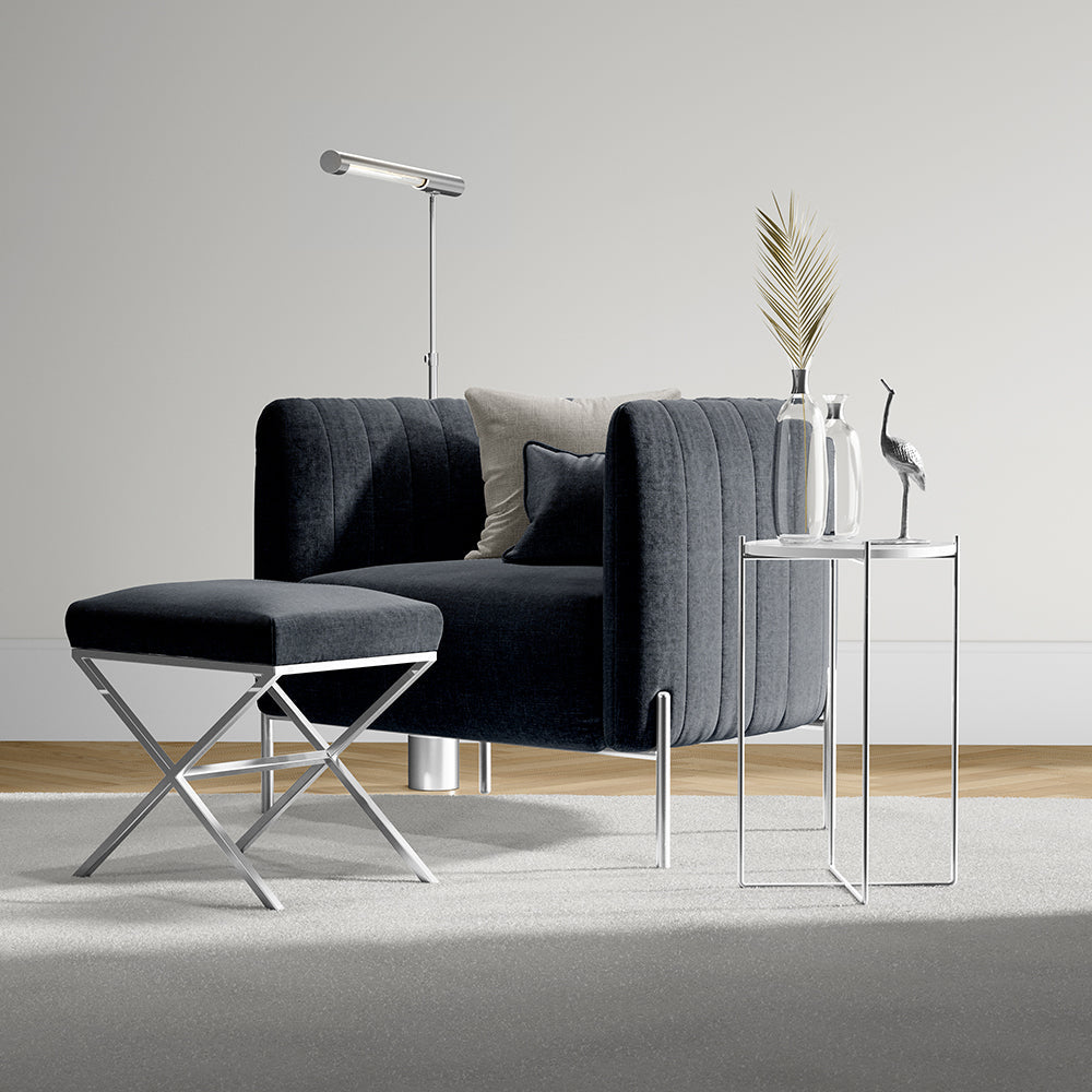 Velvet dark gray sofa with beige and navy cushions, chrome X-legs ottoman, glass-top side tables, and slim floor lamp.