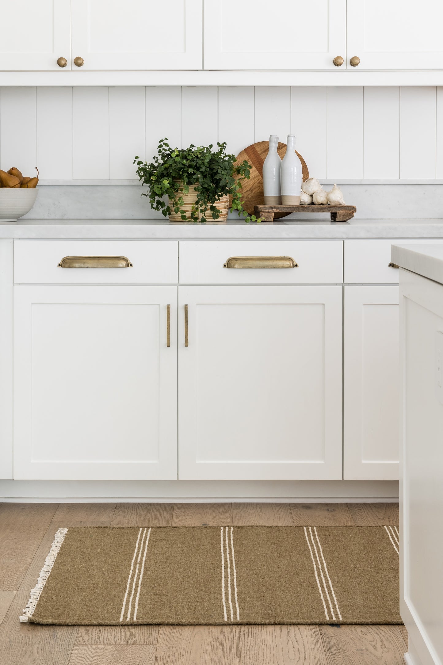 White kitchen with brass handles, marble countertop, plant in wicker basket, bottles and garlic on board, beige striped rug.