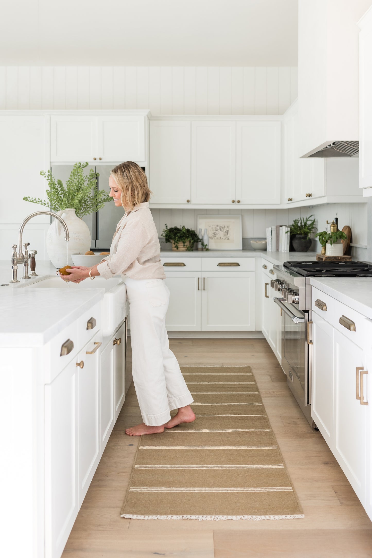 Bright white kitchen with white cabinets, brass hardware & stainless range; barefoot woman at sink, beige striped runner rug.