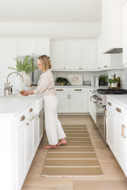 Bright white kitchen with white cabinets, brass hardware & stainless range; barefoot woman at sink, beige striped runner rug.