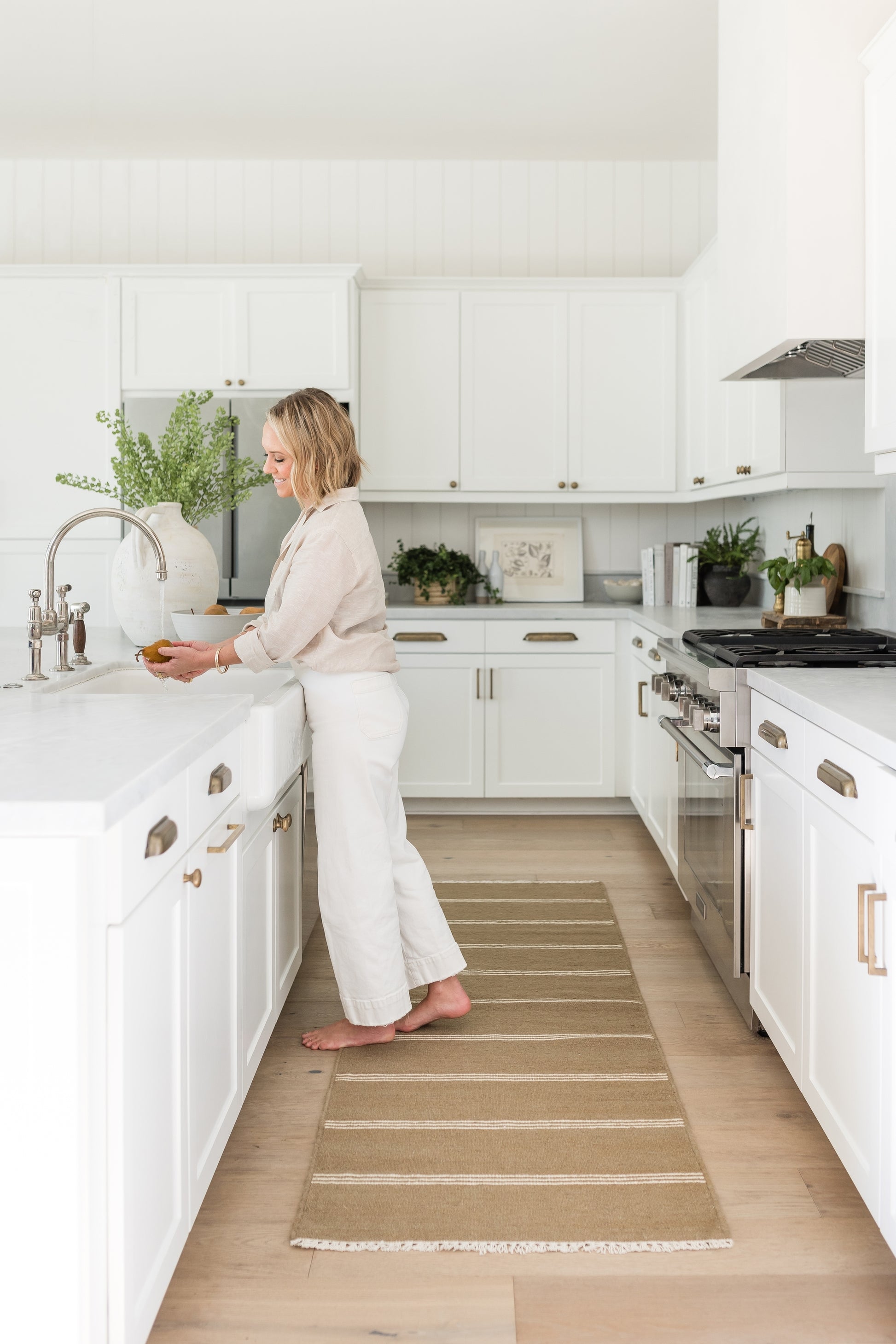 White modern kitchen with shaker cabinets and stainless range; tan striped runner rug (6' x 9'); greenery; barefoot woman at island.