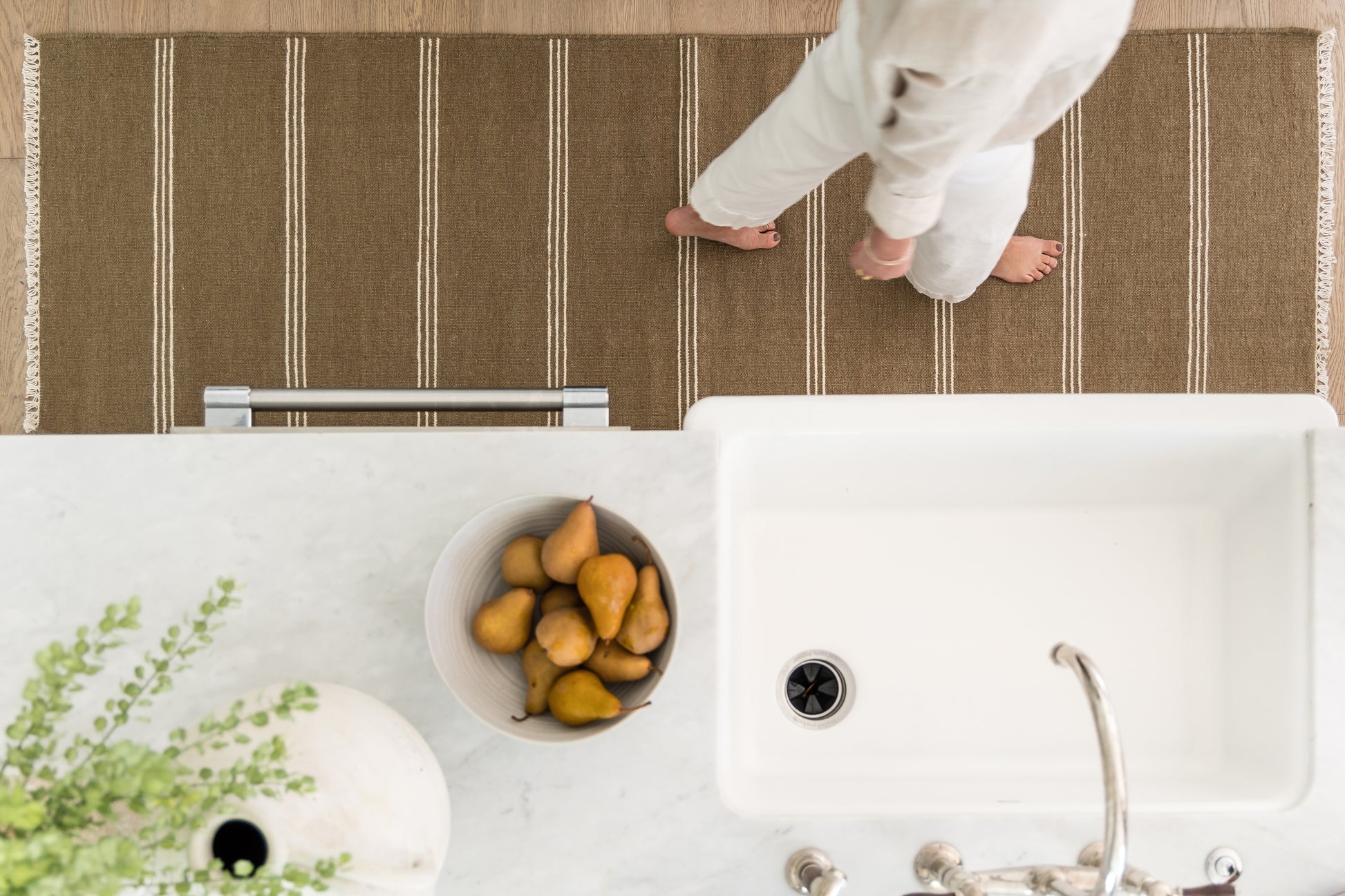 Overhead kitchen scene: marble counter, white sink, bowl of pears, green plant, brown striped runner rug, barefoot person walking