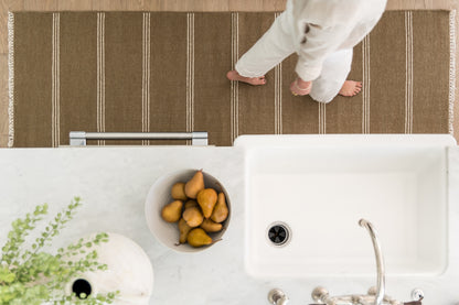Olive-brown woven rug with ivory vertical stripes and fringed ends on a wooden floor.
