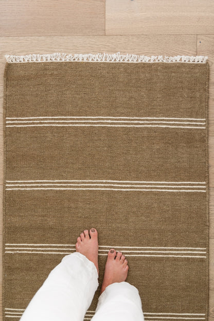 Brown jute rug with white stripes and fringe along top edge, rectangular on wood floor, feet for scale.