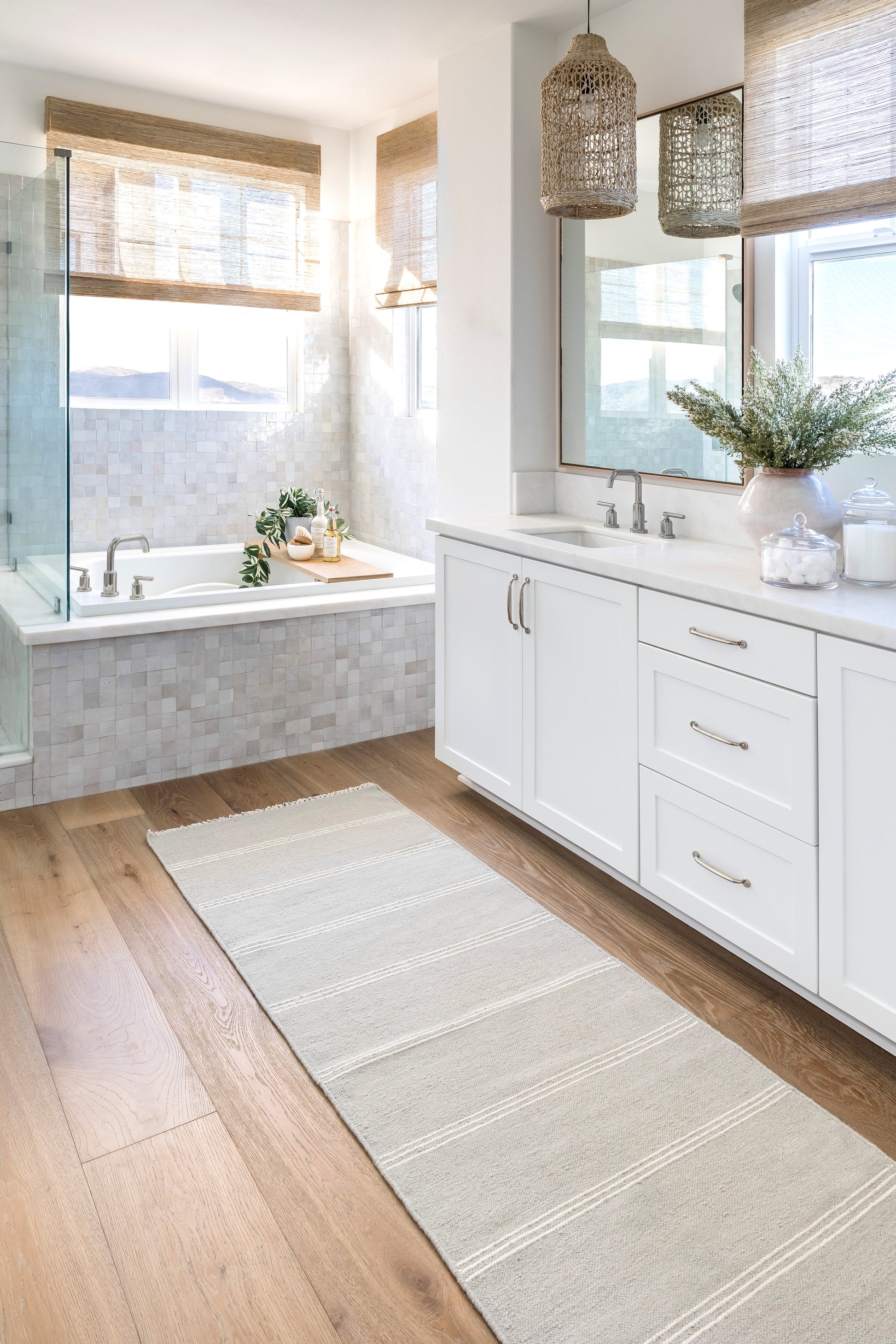 Beige woven rug on a bright coastal bathroom floor, with white cabinets, marble counters, a tub, glass shower and woven pendant lights.