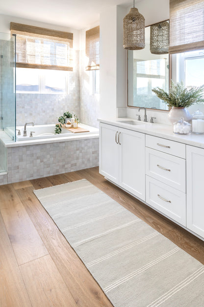 Beige woven rug on a bright coastal bathroom floor, with white cabinets, marble counters, a tub, glass shower and woven pendant lights.