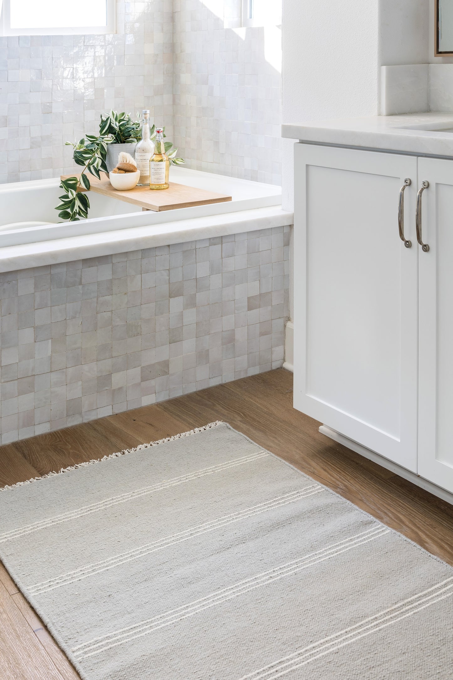 Bright bathroom with grey mosaic tub tiles, wood bath tray holding a plant and bottles, white cabinet, grey striped rug.