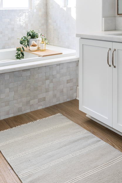Bright bathroom with grey mosaic tub tiles, wood bath tray holding a plant and bottles, white cabinet, grey striped rug.