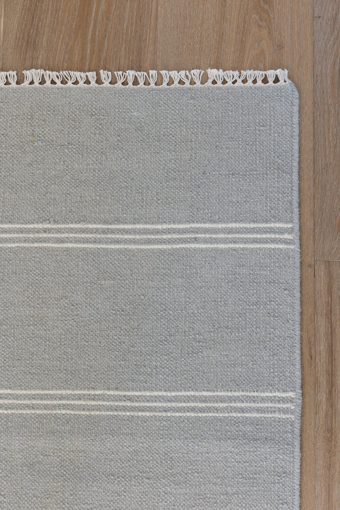 Gray woven runner rug with white fringe along the top edge and two white stripes across, on a wooden floor.