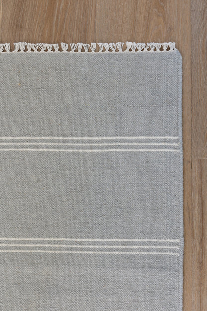 Gray rectangular area rug with two white stripes and fringe trim along the top edge on a wood floor.