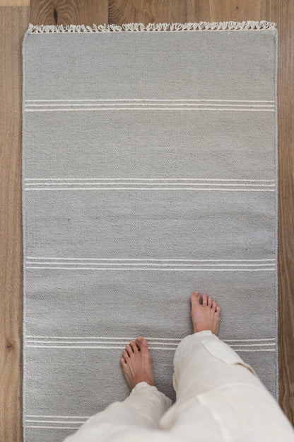 Gray woven runner rug with white horizontal stripes and fringe at top, placed on wood floor; barefoot person standing on it.