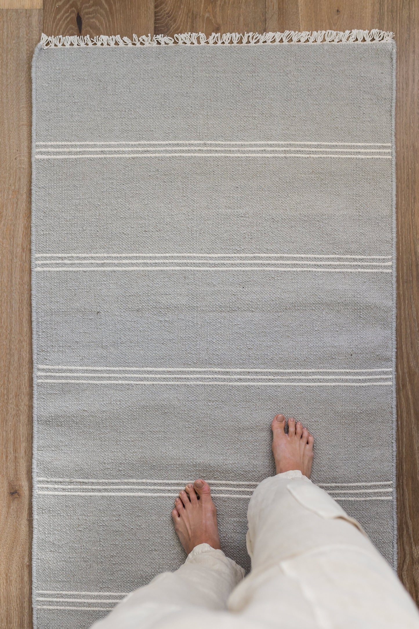 Gray woven rug with white stripes and fringe laid on wood floor, seen from above with bare feet in light pants.