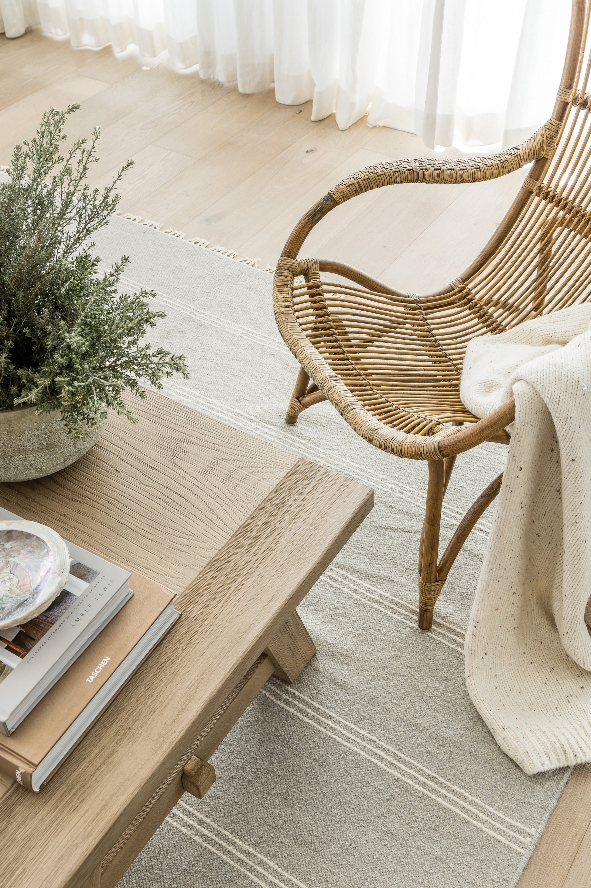 Woven rattan lounge chair beside a light wood coffee table, potted greenery, cream rug, sheer white curtains.
