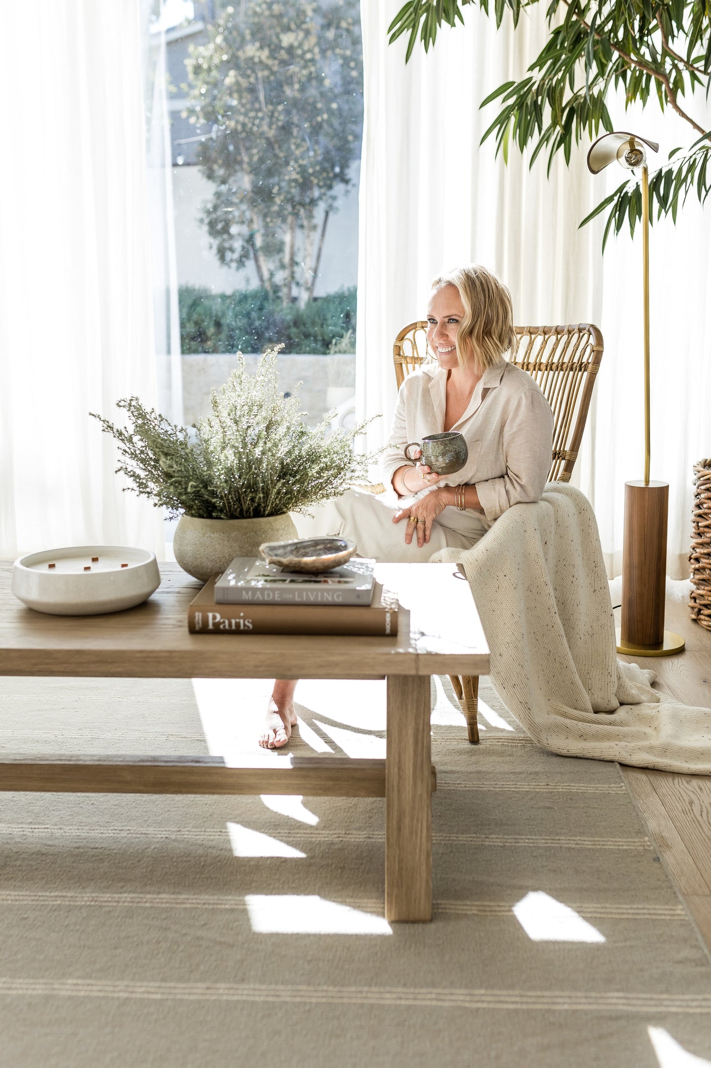 Bright living room with a beige 3' x 8' runner rug, wicker chair, wood coffee table, potted greens, books, and a person holding a mug.
