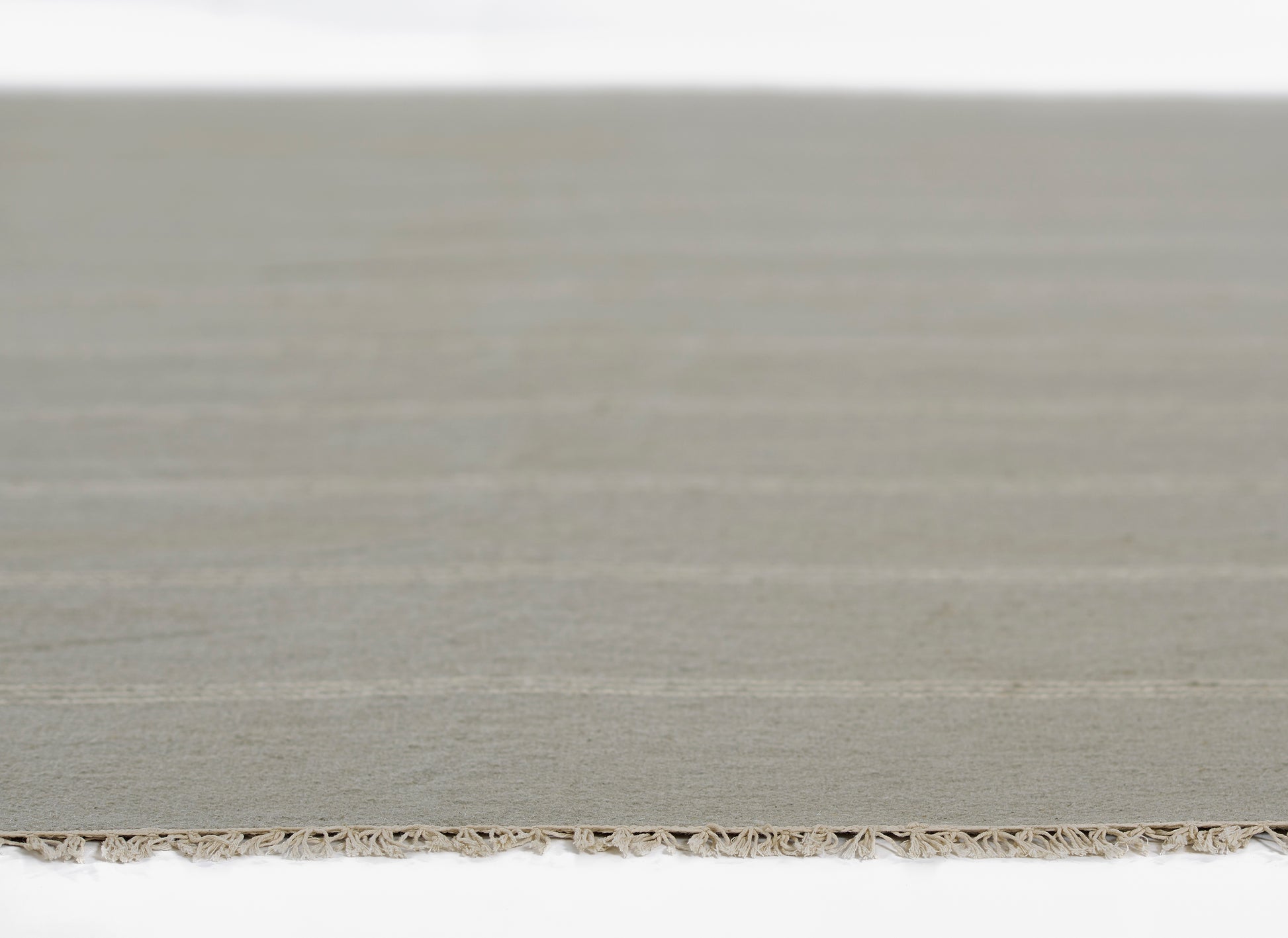 Neutral beige-gray flat-weave rug with a fringe along the bottom edge.