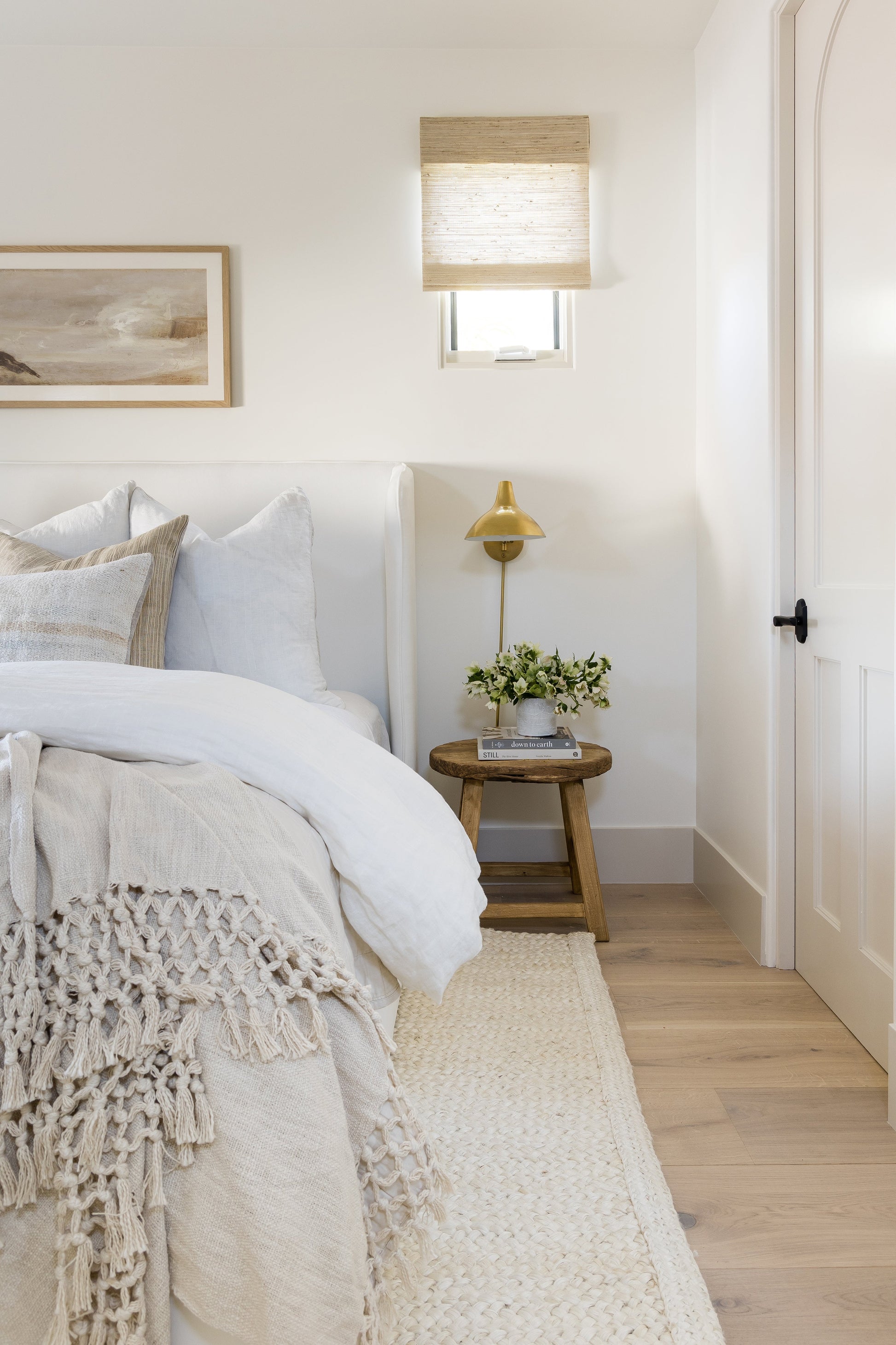 Neutral rug on the floor in a bedroom setting, with white linen bedding, beige knitted throw, round wooden side table, brass lamp, greenery and art.