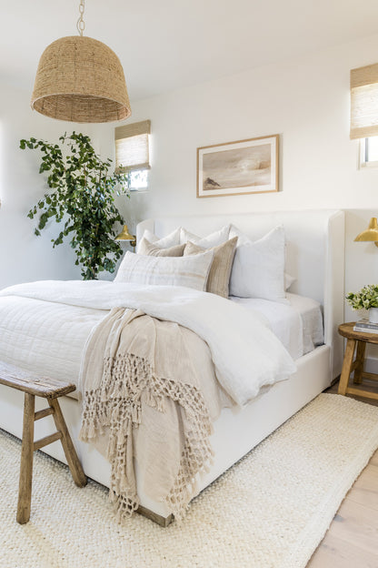 Bright neutral bedroom with white upholstered bed, beige knit throw, layered pillows, wicker pendant lamp, plant, rug & bench.