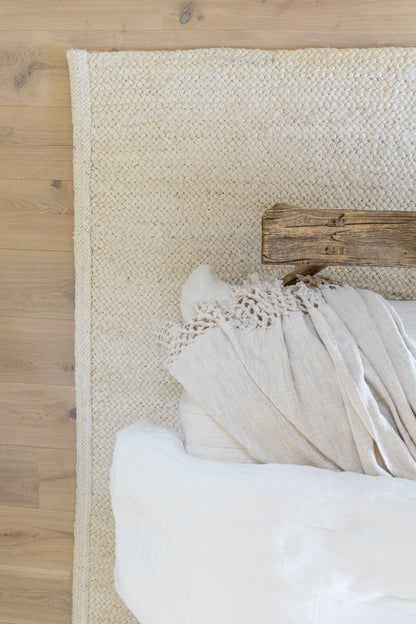 Cream braided jute rug on light wood floor with beige linen throw and white cushions, rustic cozy living room decor.