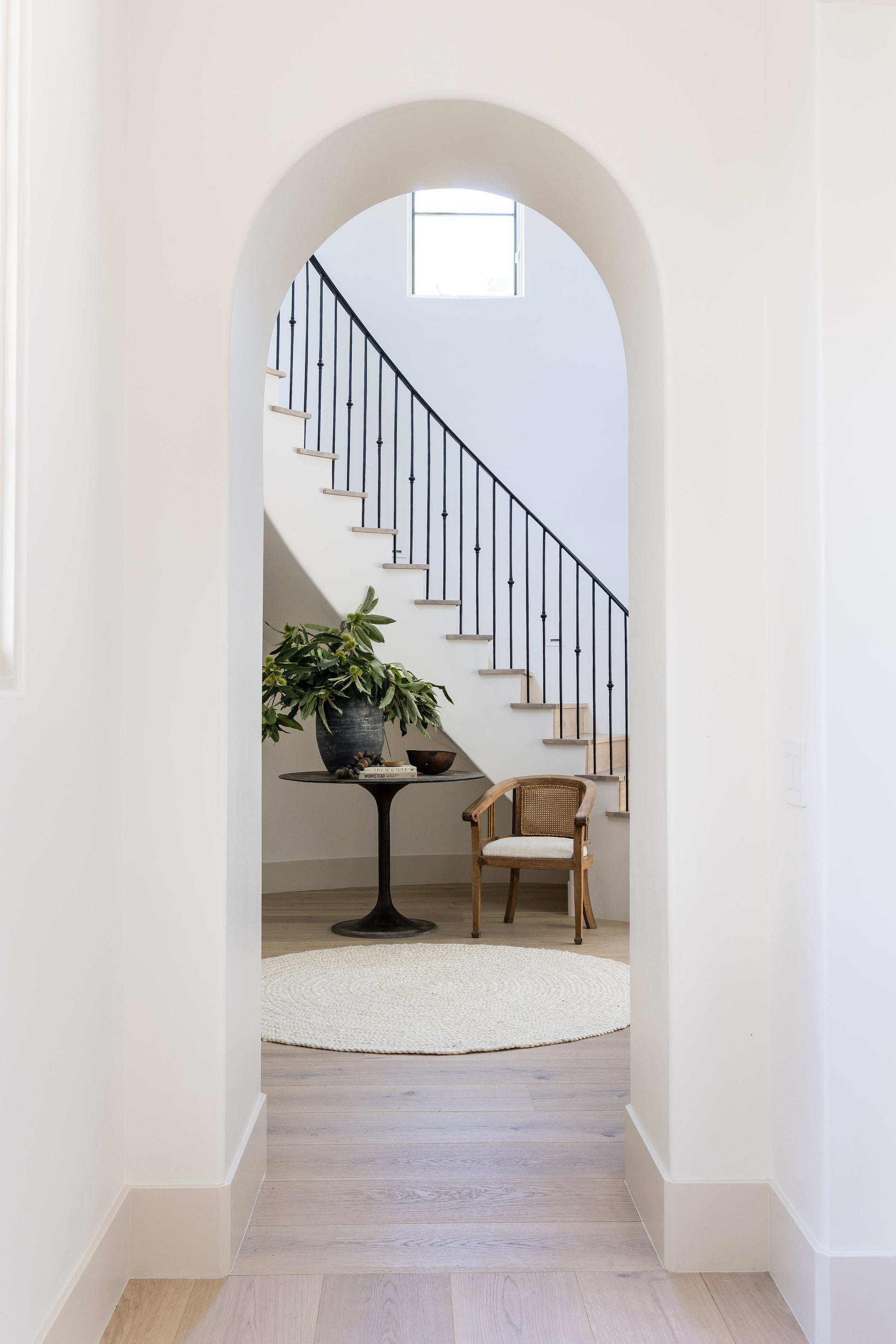 Arched entryway to a cozy foyer: rectangular rug, pedestal table with plant, wicker chair, and black stair railing.