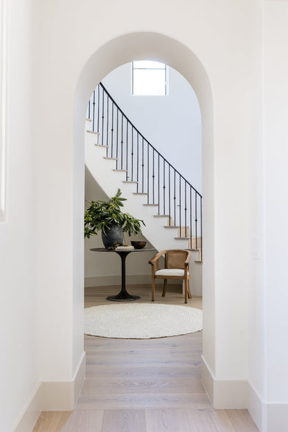 Arched entryway to a cozy foyer: rectangular rug, pedestal table with plant, wicker chair, and black stair railing.