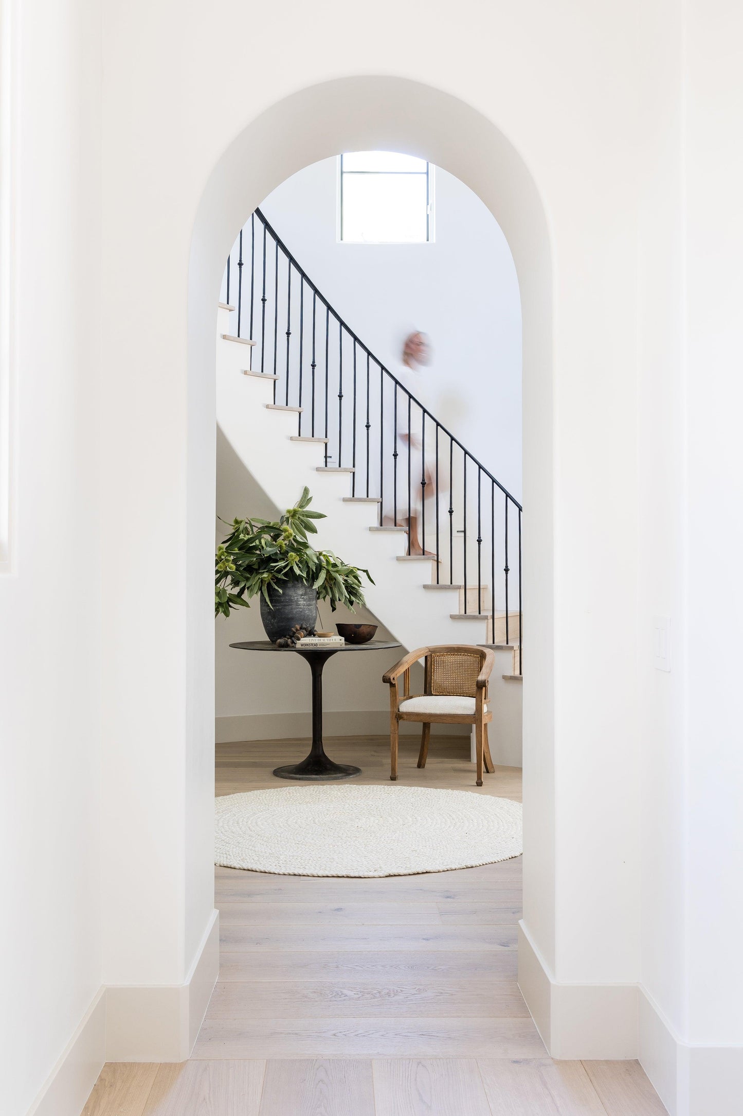 Bright entryway with an arched doorway into a stair hall; rug visible on the floor near a black railing, a wicker chair, and a round pedestal table with a plant.