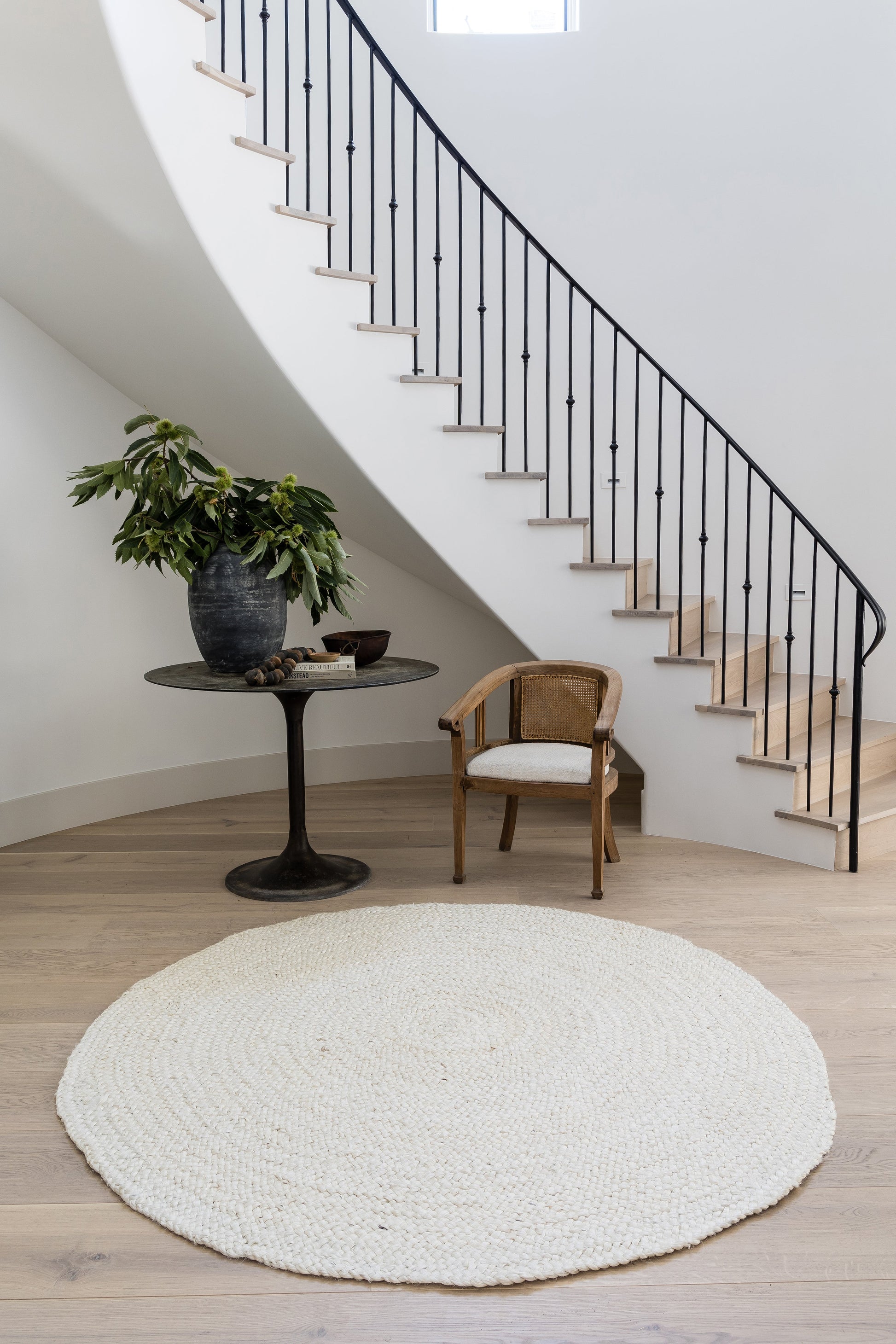 Curved staircase with black metal balusters, cream rug, wooden floor, potted plant on pedestal table, wicker chair.