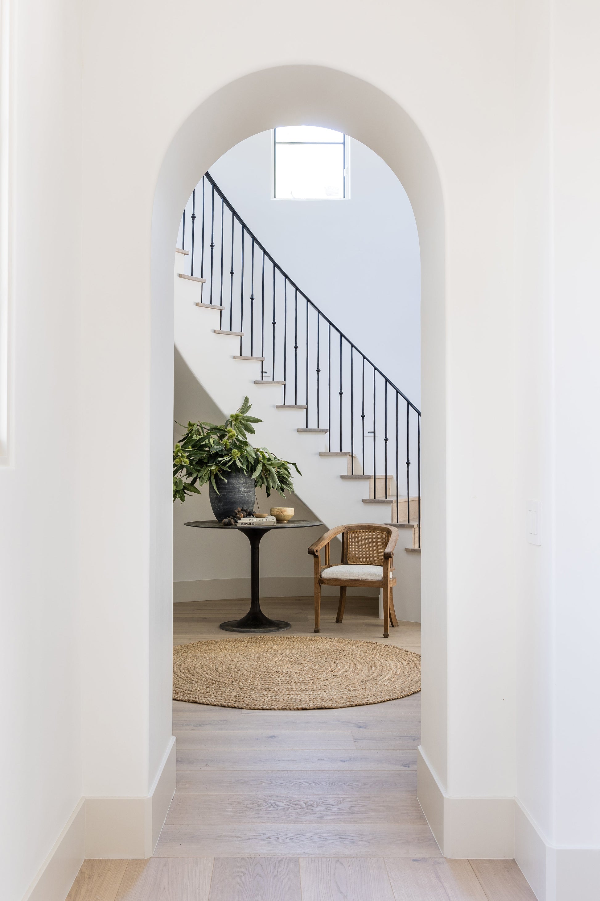 White archway to a stair with black railing, round table with plant, woven chair, and circular jute rug.