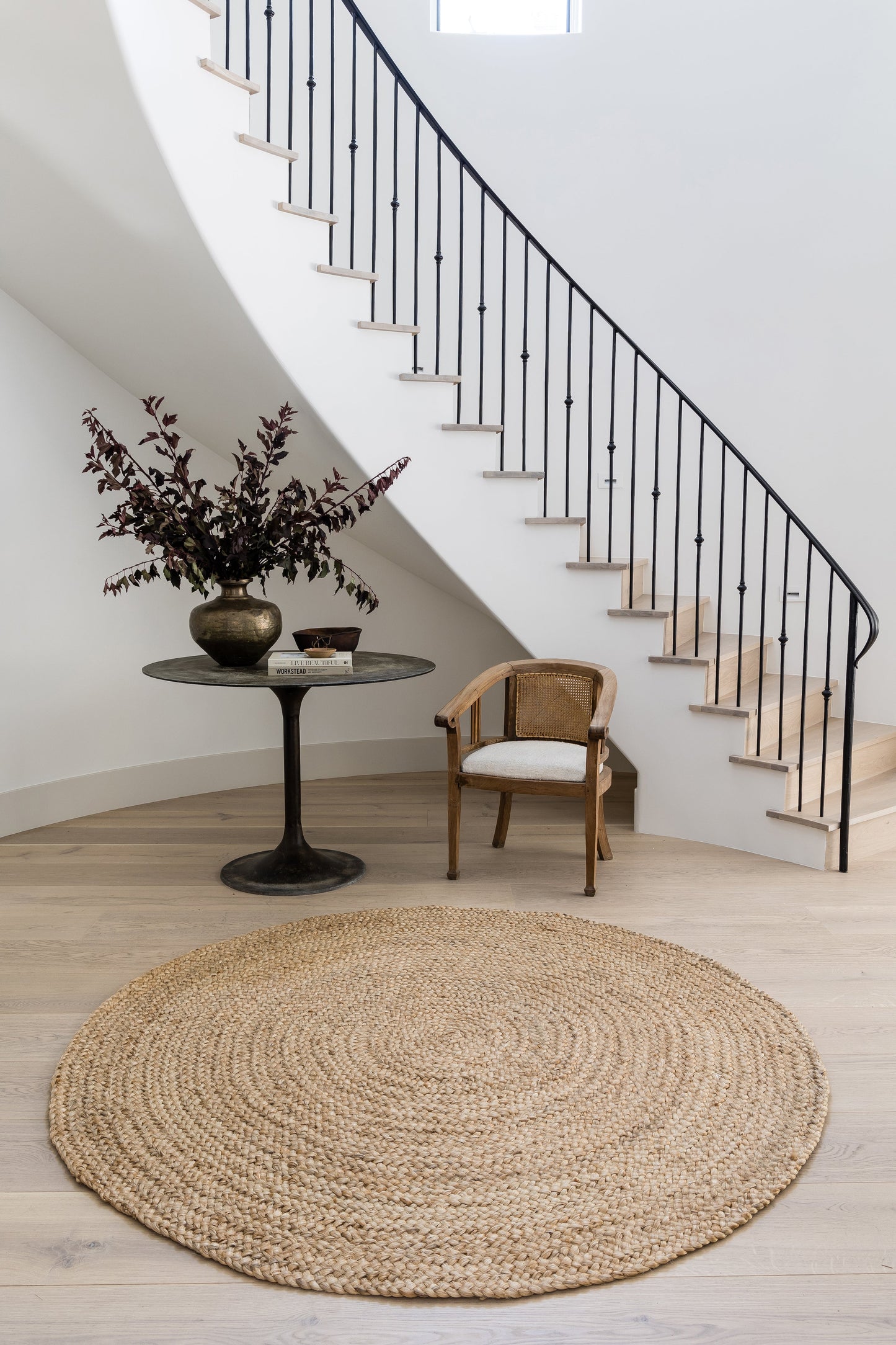 Natural round jute rug in a bright entry with a black pedestal table, vase of dark foliage, and woven armchair.