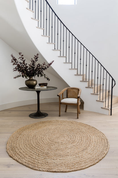 Natural round jute rug in a bright entry with a black pedestal table, vase of dark foliage, and woven armchair.
