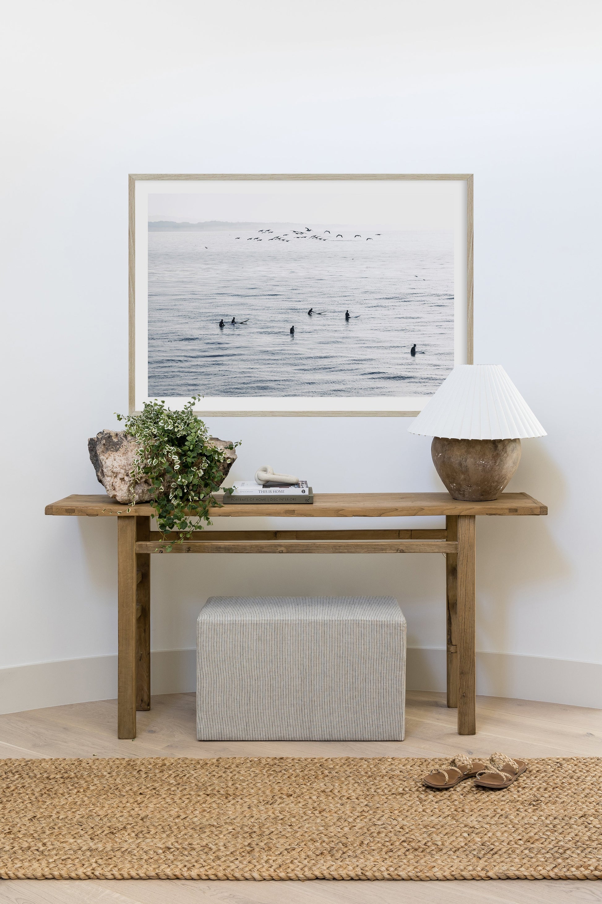Wooden console table with framed seascape, potted greenery, books, beige lamp, gray striped ottoman, jute rug.