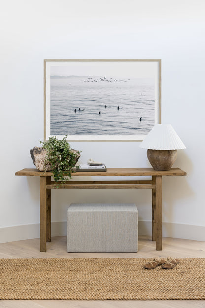 Natural wood console table with green plant, framed ocean photo, seashell accents, ceramic lamp, striped ottoman, jute rug.