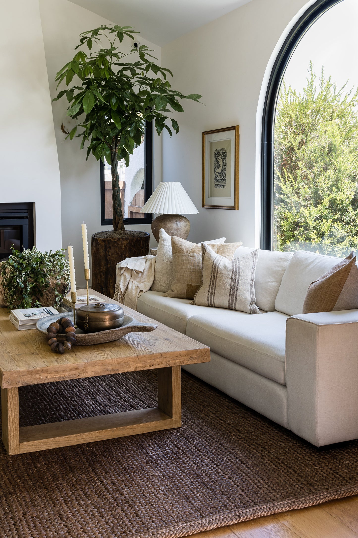 Neutral living room with a 3' x 8' rug runner on the floor, white sofa with beige striped cushions, tall potted tree, rustic wood coffee table, arched window.