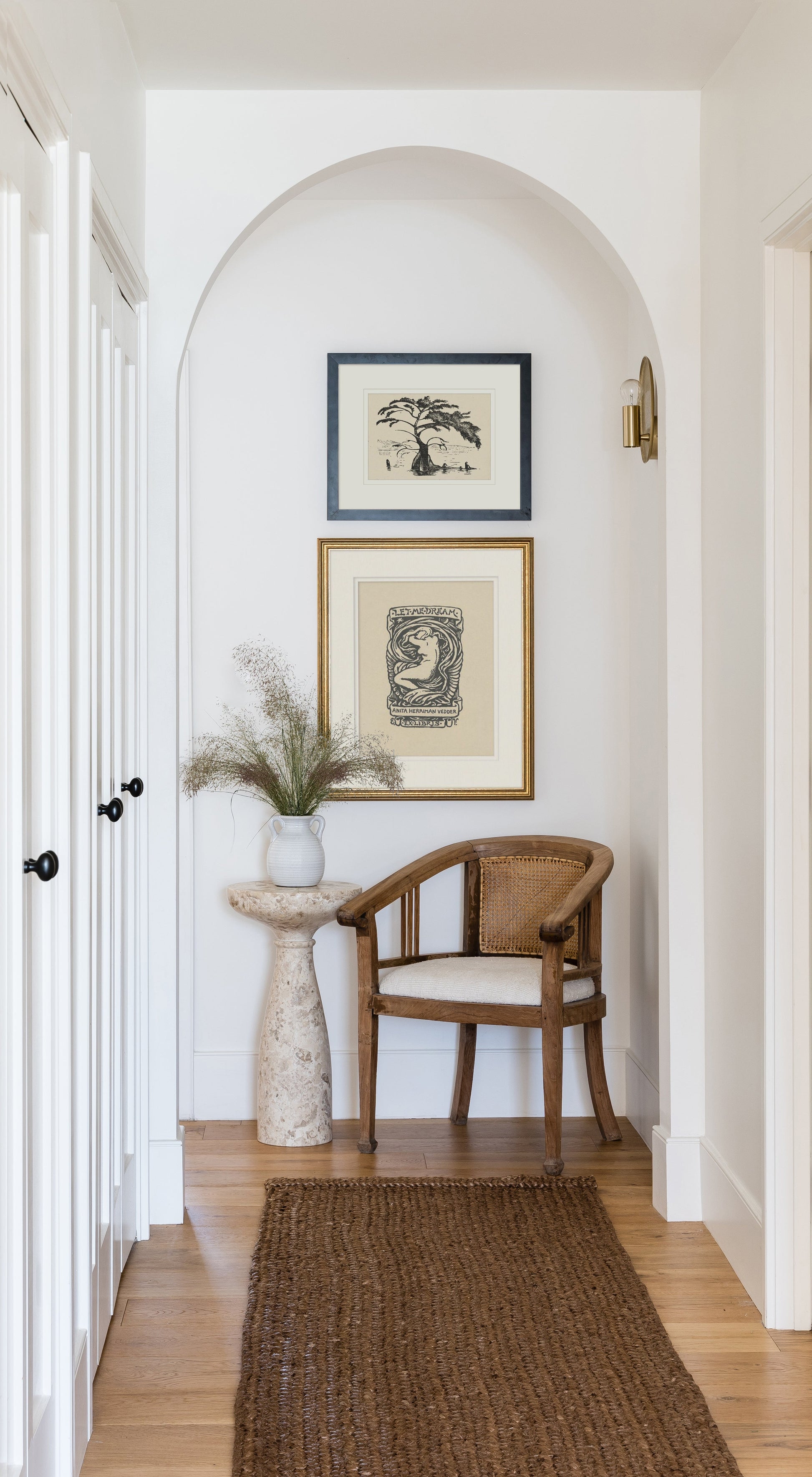 White hallway alcove with arch, framed prints, wooden cane chair, stone pedestal vase with dried grasses, brown woven rug.