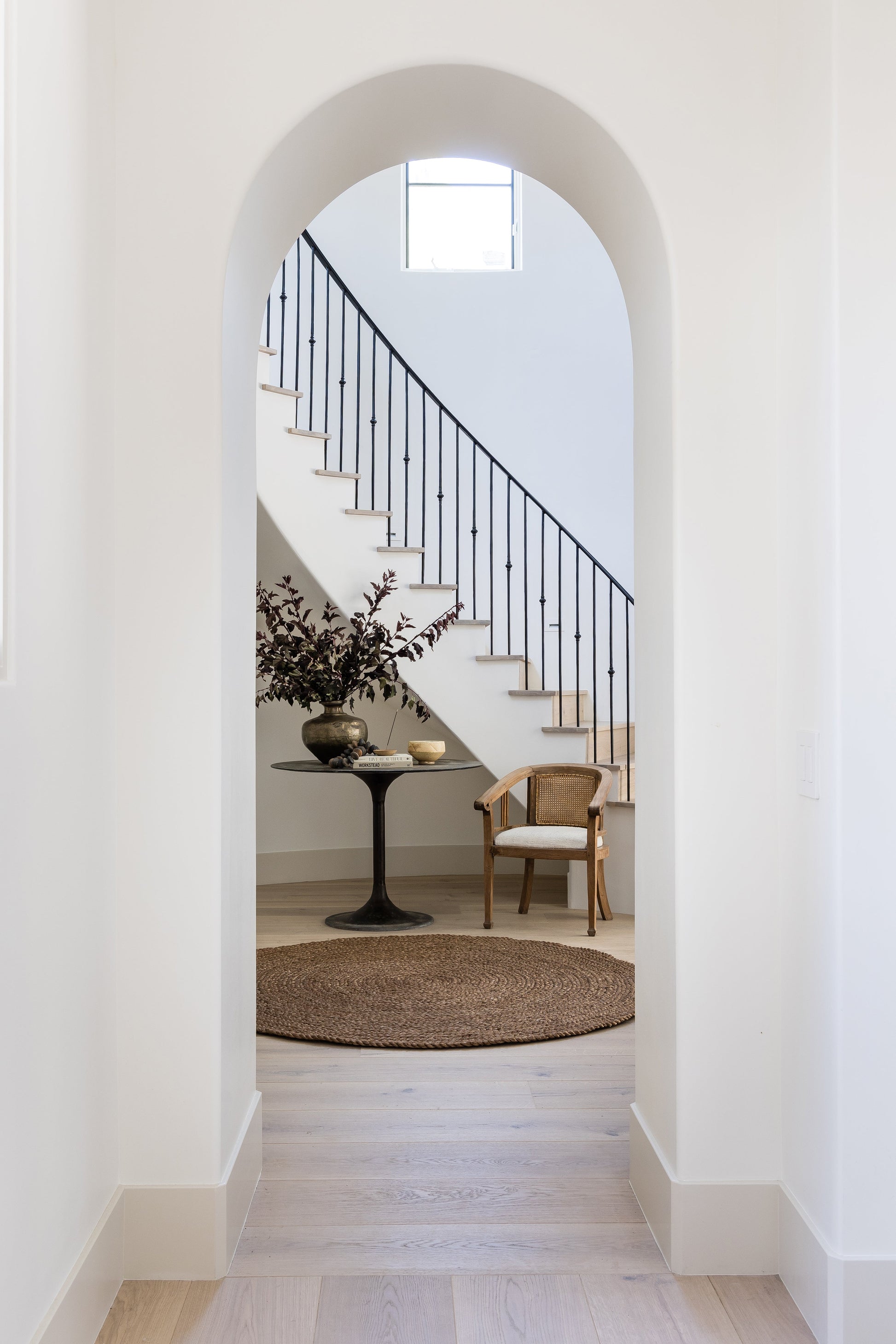 Arched white hallway view of staircase with black iron railing, woven rug, pedestal table with vase, and a wicker chair.