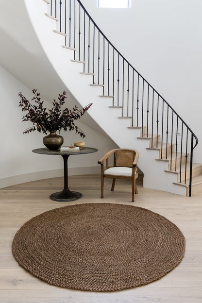 Round natural jute rug on light wood floor, black pedestal table with brass vase and books, cane chair under curved stairs.