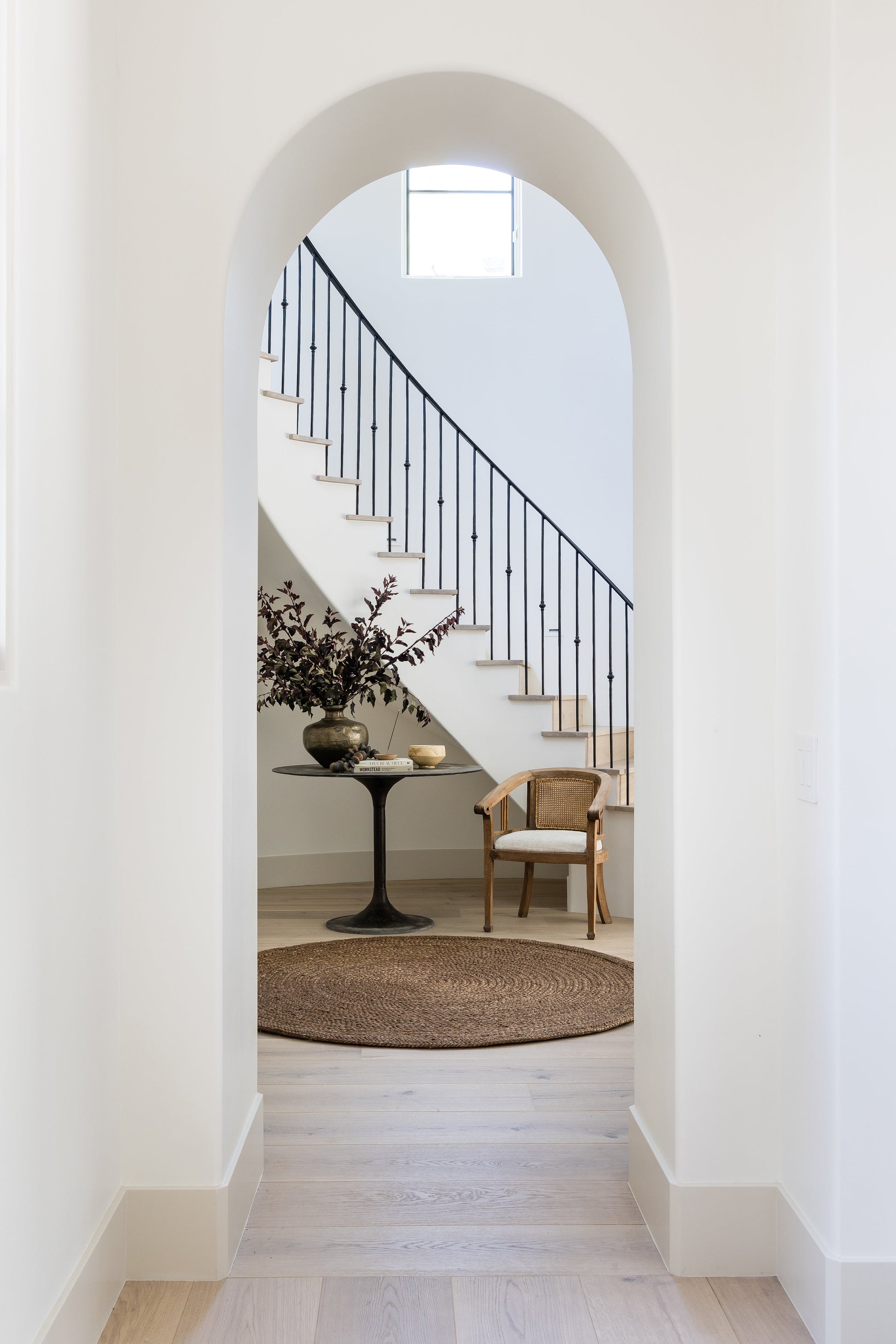 White arch doorway into a living area with black balustrade staircase, round jute rug, pedestal table, vase, and cane chair.