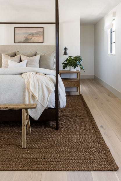 Neutral bedroom with dark wood four-poster bed, gray bedding, beige throw, jute rug, rustic wooden bench, and potted plant.