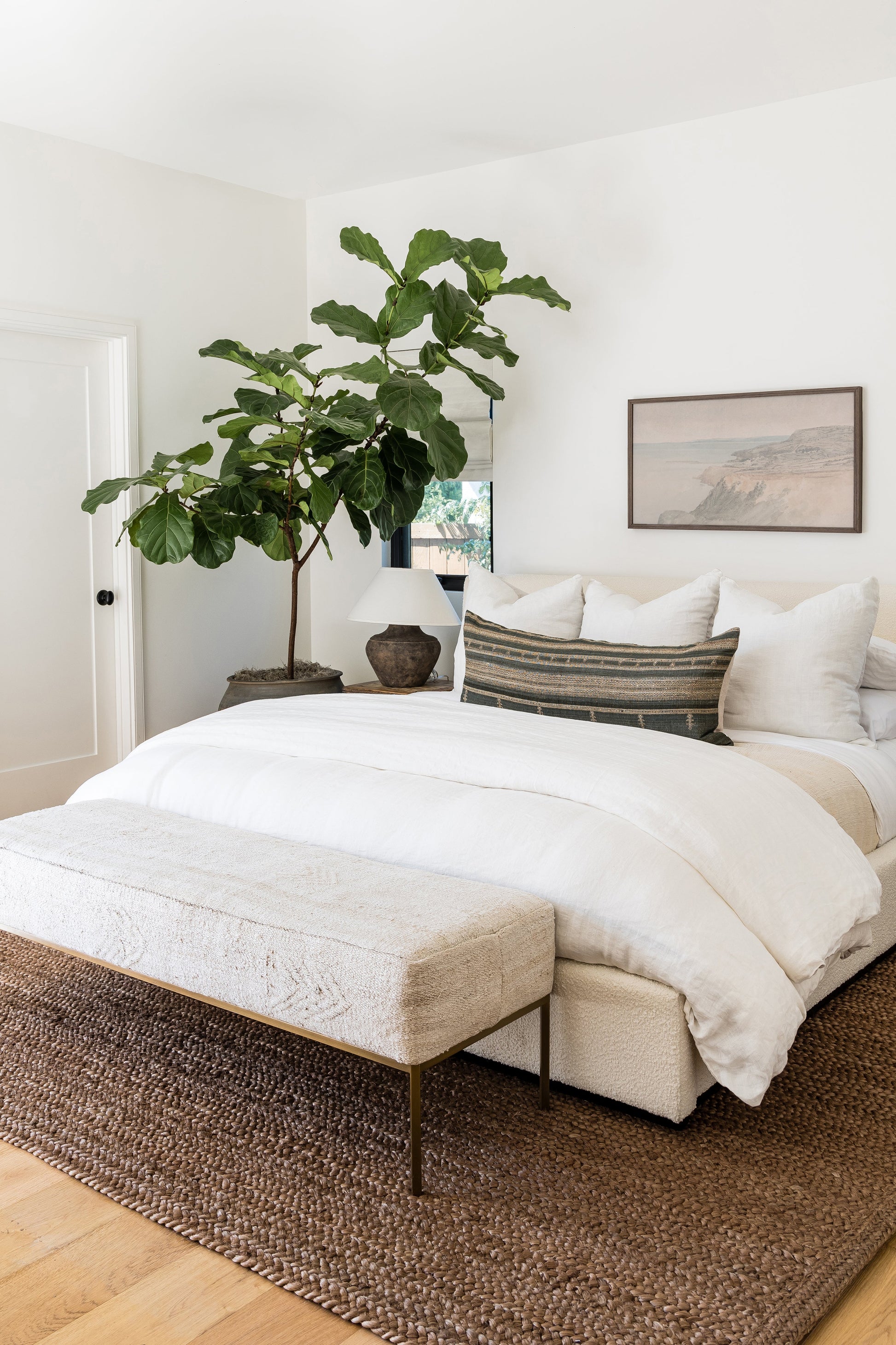 Neutral bedroom scene with a woven jute rug on the floor, white bedding on a low bed, a beige upholstered bench, and a large fiddle-leaf fig plant.