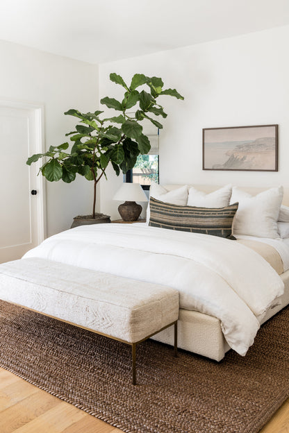 Tall fiddle-leaf fig in a brown pot beside a white duvet bed with striped lumbar pillow and cream bench on a round woven rug.