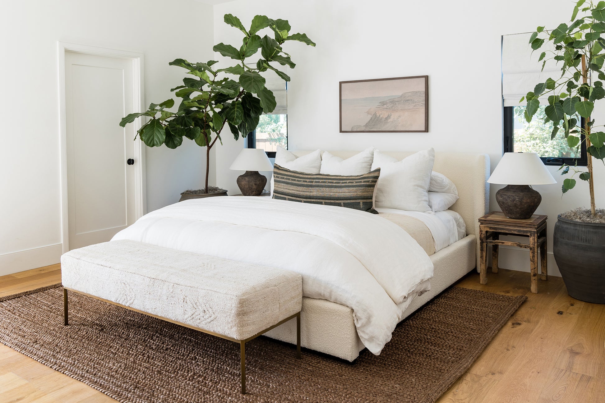 Cream upholstered bed with white duvet, beige foot bench, brown jute rug, two nightstands with lamps, large potted plants.